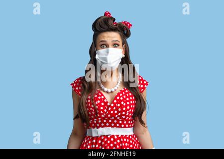Shocked pinup lady in retro clothes wearing face mask against coronavirus on blue studio background Stock Photo