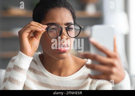 Astigmatism. Young Black Female In Eyeglasses Trying To Read Message On Smartphone Stock Photo
