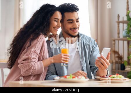 Happy Middle Eastern Couple Browsing Internet On Smartphone While Having Breakfast Stock Photo