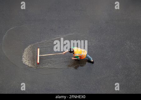 Worker cleans the street after the rain. Janitor with brush washes wet city road, view from the top Stock Photo