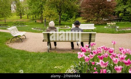A senior couple, woman and man wearing a hat, sitting on a white bench in the park among flowers. Stock Photo