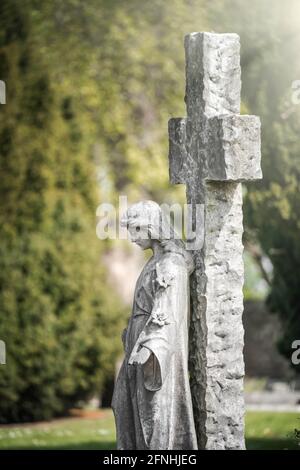 Stone old weathered grave statue of granite weeping angel with hand held out.  Standing against large stone crucifix cross sad peaceful in beautiful Stock Photo