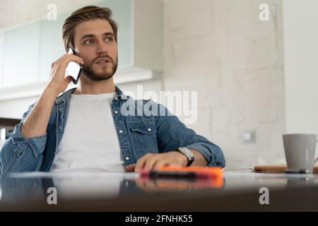 Smiling man, he is sitting at a table, talking on the phone with his laptop, notebook in front of him Stock Photo