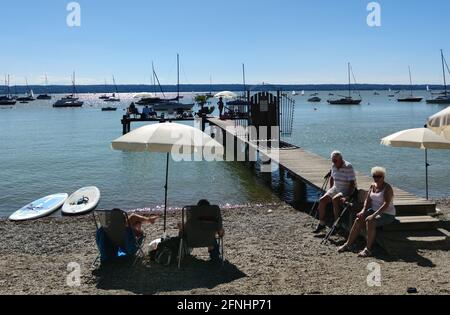 An elderly couple dressed in summer clothes sit on the jetty of a sandy beach of a lake and rest. The man has two crutches. Stock Photo
