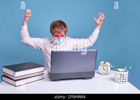 Angry man in medical face mask works behind laptop on blue studio background Stock Photo