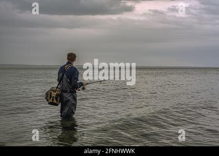 young man stands alone in waders fishing with a casting rod in the gloomy morning, Denmark, May 9, 2021 Stock Photo