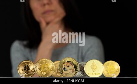 Variety of trending Symbolic cryptocurrency types represented as shiny coins are lined up against dark background. A woman with her hand on chin is un Stock Photo