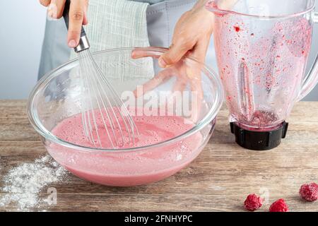 A caucasian woman is mixing a red fruit juice made with fresh berries in a glass bowl using a balloon whisk. Versatile food preparation image for home Stock Photo