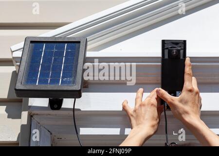 A caucasian woman is installing a wireless security surveillence camera with motion sensor on top of the front door frame. The camera is linked to a s Stock Photo