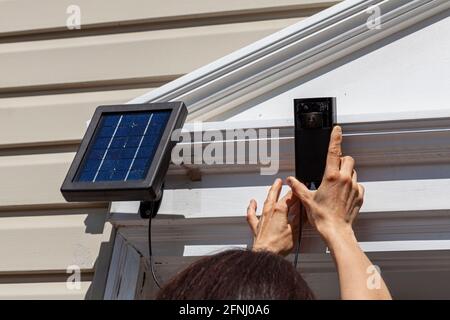 A caucasian woman is installing a wireless security surveillence camera with motion sensor on top of the front door frame. The camera is linked to a s Stock Photo