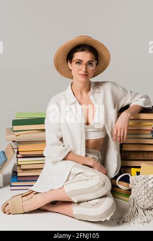 pleased woman in straw hat and glasses sitting near stack of books on white Stock Photo
