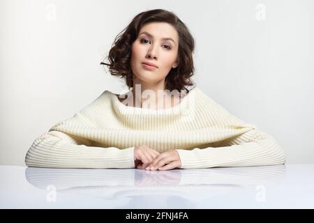 Beautiful brunette woman dressed in large white woolen sweater is sitting at white table on gray background. Stock Photo