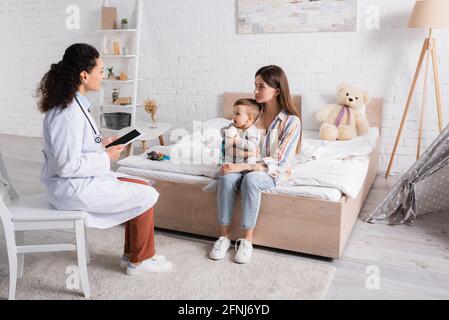 african american pediatrician in white coat holding digital tablet with blank screen near mother and son in bedroom Stock Photo