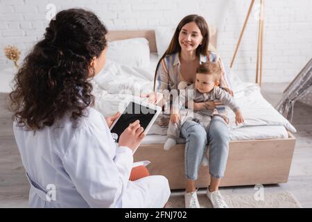 african american pediatrician in white coat using digital tablet with blank screen near happy mother and son in bedroom Stock Photo
