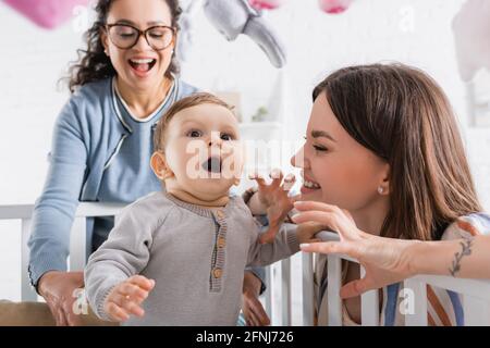smiling mother gesturing near amazed infant son in baby crib and excited african american friend Stock Photo