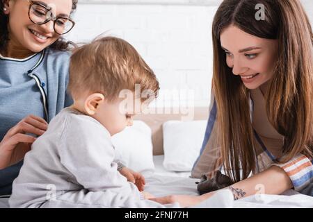 interracial women looking at infant boy sitting on bed Stock Photo
