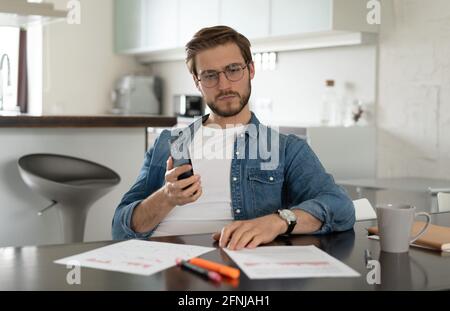 Confident man sitting at a table holding smartphone, chatting online with colleagues, looking at documents Stock Photo