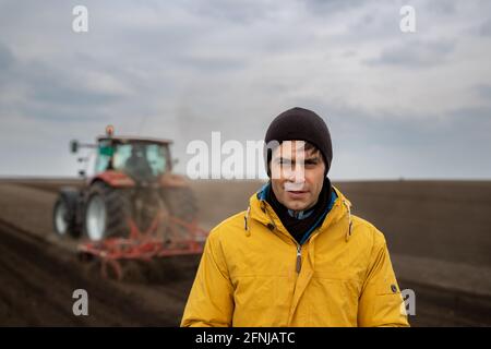 Portrait of handsome farmer standing in front of tractor harrowing in field in background Stock Photo