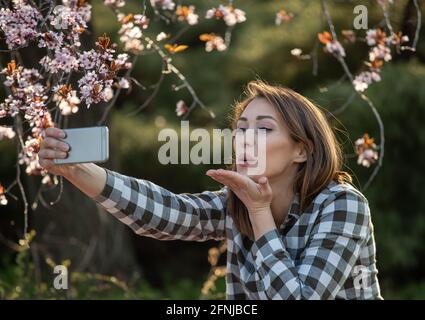 Happy young girl sending kisses over mobile phone in front of blooming fruit tree in spring time Stock Photo