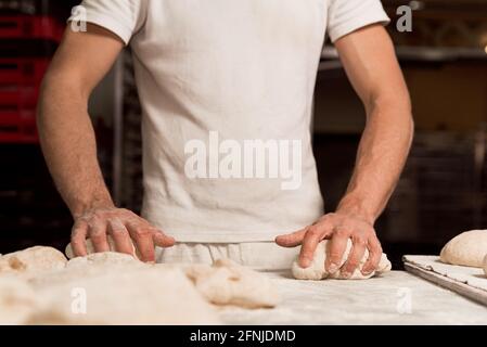 young male baker in bakery factory manually kneading fresh bread knead with flour on worktop . concept of traditional manual bread preparation in brea Stock Photo