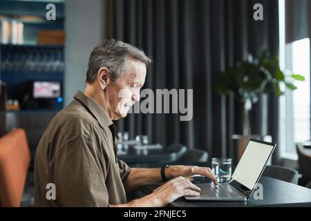 Side view of serious busy senior freelancer with mustache sitting at black table in cafe and performing task on laptop Stock Photo