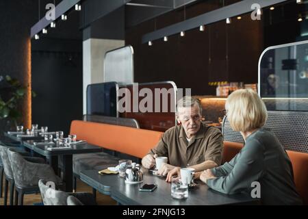 Surprised senior Caucasian man with mustache holding tea cup while chatting with woman in modern fashionable cafe Stock Photo