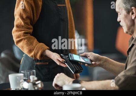 Serious senior man sitting at table and using NFC on smartphone while paying for lunch in cafe, waitress in apron holding payment terminal Stock Photo
