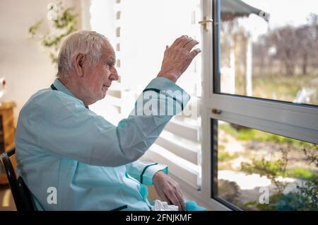 Senior man sitting in wheelchair, looking through big window and waving hand for greeting. Social distancing concept Stock Photo