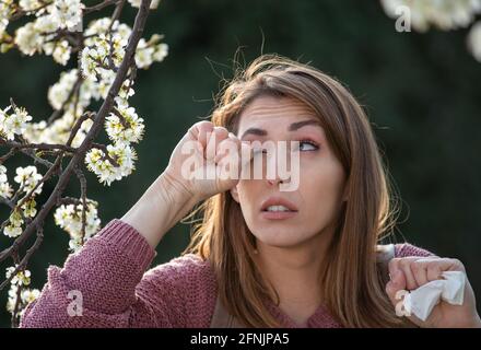 Young pretty girl rubbing eyes beside blooming tree in spring time. Itchy eyes from allergy Stock Photo