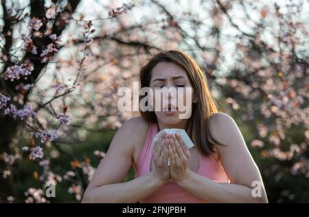 Pretty young woman sneezing in front of blooming tree. Spring allergy attack concept