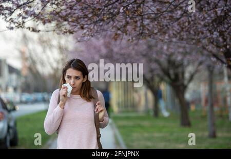 Pretty young girl walking in street under blooming tree in springtime and having allergy reaction on pollen of runny nose Stock Photo