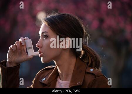 Pretty young woman using asthma pump in front of blooming tree in spring. Seasonal allergy attack Stock Photo