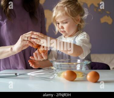 Adorable little girl with blonde hair helping mom to cook pie, mother showing daughter how to break fresh eggs into bowl while cooking together in kitchen at home, family preparing food on weekend Stock Photo