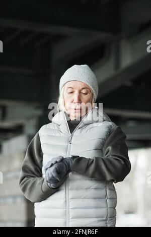 Serious senior Caucasian woman in gray hat and gloves standing under bridge and checking pulse on tracker after running Stock Photo