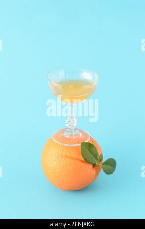 A glass filled with orange juice stands on an orange that also has green leaves on a blue background. A summer refreshing citrus drink with a creative Stock Photo