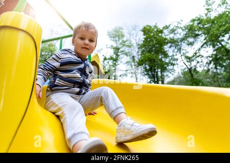 Portrait of cute little cute caucasain blond toddler boy enjoy having fun sliding down modern plastic slide at outdoors playground area city street Stock Photo