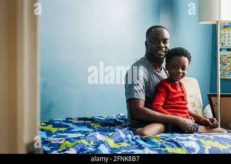 Portrait of cute boy sitting with father on bed against wall at home Stock Photo