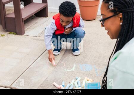 Boy using chalk to draw on concrete while mother smiles and helps Stock Photo