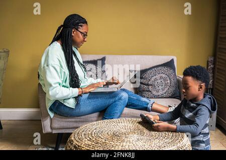 Mother using laptop while son playing game on mobile phone in living room at home Stock Photo