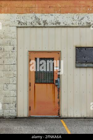 Orange security door against white and orange brick wall and cream-coloured siding. Keyless numbered door padlock and bars on window. Boarded window. Stock Photo