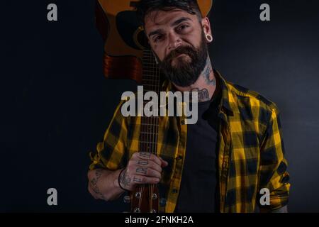 Tattooed man with a beard wearing a tartan yellow black shirt is  posing holding his guitar on his sholder Stock Photo