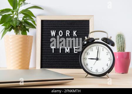 Classic vintage black alarm clock, laptop, potted plants and board sign 'work time' on wooden desk against white wall background. Time and work concep Stock Photo