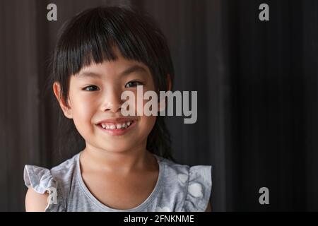 Portrait charming Asian little girl, smiling face, bang black hair, showing healthy white teeth and gum, black metal background. Space for text. Stock Photo