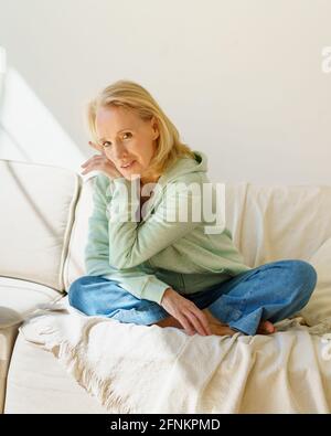 Enjoying retired life. Portrait of happy pleasant mature 60s woman in casual wear resting on coach and smiling at camera, vertical shot. Elderly female retired grandmother enjoying free time at home Stock Photo