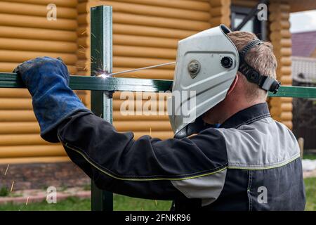Close up of a young  man welder in  uniform, welding mask and welders leathers, weld  metal  with a  welding machine at the construction  of a fence i Stock Photo