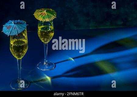 Two flute glasses with sparkling yellow liquid with colored paper umbrellas on top, cast reflections and shadows on a blue surface Stock Photo