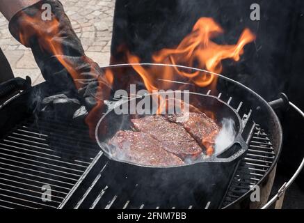 Chef searing three portions of Denver Steaks in a skillet over the flames in a close up on the pan and his gloved hand Stock Photo