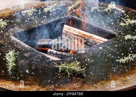 Fresh sprigs of rosemary around a barbecue fire with burning logs and smoke in a large outdoor metal griddle Stock Photo