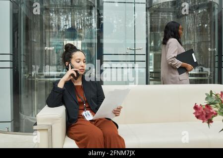 Young businesswoman sitting on sofa in the office corridor reading document and talking on mobile phone Stock Photo