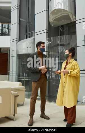 Business colleagues talking in masks while standing at office corridor Stock Photo